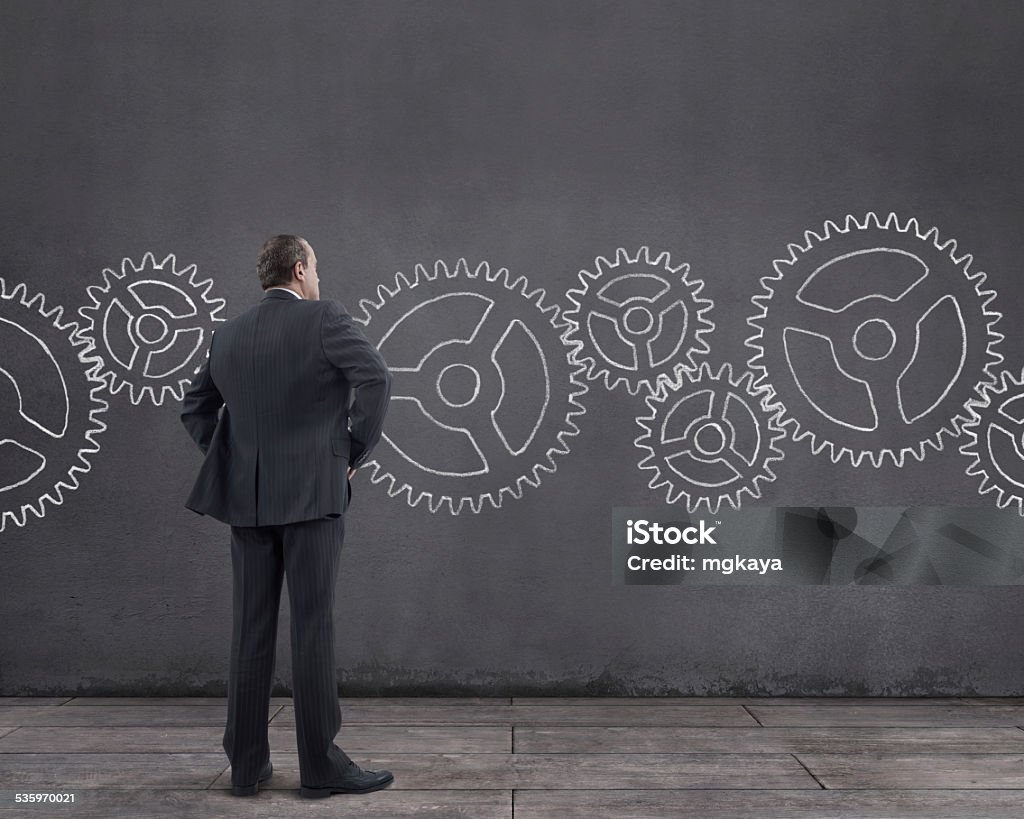 Businessman and Gears Rear view of businessman standing on wooden floor with gears sketched (chalk drawing) on the wall. He is looking further, thinking, searching new ideas. Domestic Room Stock Photo