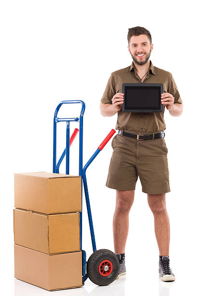Messenger showing a digital tablet Smiling courier standing close to push cart and showing a digital tablet. Full length studio shot isolated on white. mover delivery person box hand truck stock pictures, royalty-free photos & images