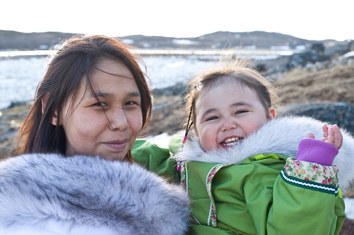 An Inuit mother and her daughter wearing traditional amouti on the remote tundra of Baffin Island in late spring.  They are interacting with each other, smiling and laughing.  Background is barren land of rock, ice and distant mountains.