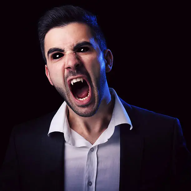 Scary man in suit with black eyes and fangs screaming against black background.