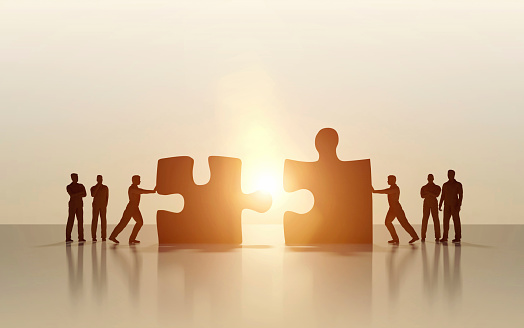 Group of business men supporting two of them pushing jigsaw puzzle pieces together, creating connection as a result of successful teamwork, partnership and finding solutions. Brightly lit image with dark silohuettes on a background illuminated by sunlight with lens flares. Clean and modern, empty set with copy space. Digital generated image and characters.