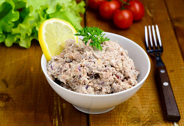 Close up appetizer: creamy tuna salad in white bowl garnished with slice of lemon and parsley leaf on wooden table with cherry tomatoes and salat