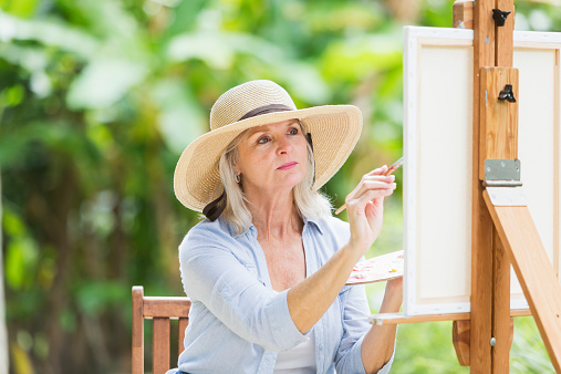 A female artist sitting outdoors at an easel, painting a picture of the landscape.  This senior woman has a serious expression on her face, staring intently at the canvas, holding a palette in one hand and a paintbrush in the other.  She is wearing a wide brimmed hat.  The background of green, lush foliage is out of focus.