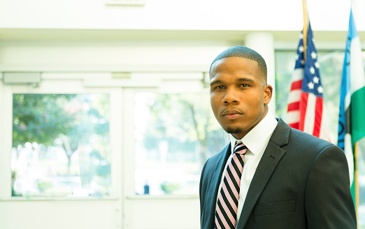 African descent, male attorney or politician in American courthouse building or congressional offices.  Or principal, professor in school building.  Red, white, and blue USA flag in background. He wears a full suit and tie and looks seriously at camera. Handsome, confident, successful.