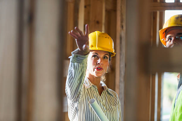Mature workers talking at construction site Two workers talking at a construction site, standing in a building in the framing stage.  The focus is on the mature woman, wearing a yellow hard hat, holding a set of rolled-up plans, pointing upward.  The male worker is cropped and partly obsured by the wooden frame. foreman stock pictures, royalty-free photos & images