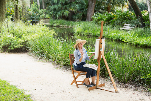 An artist outdoors by a stream sitting at an easel, painting a picture of the landscape.  She is a senior woman, relaxed and smiling as she works on her canvas.  She is dressed casually, in jeans and a wide brimmed hat.