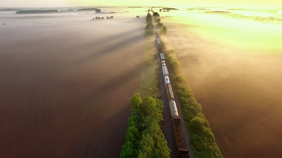 Freight train rolls across surreal, foggy landscape at sunrise, aerial view.