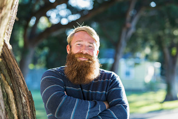 Mid adult man with a long beard Handsome man with blond hair and a long red beard, standing outdoors on a sunny day.  He is smiling, looking at the camera with his arms folded, standing next to a tree. bushy stock pictures, royalty-free photos & images