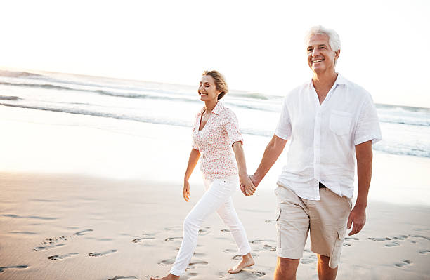 We may not be newlyweds but we act like it A senior couple walking hand-in-hand on the beach couple holding hands while walking on beach at sunset stock pictures, royalty-free photos & images