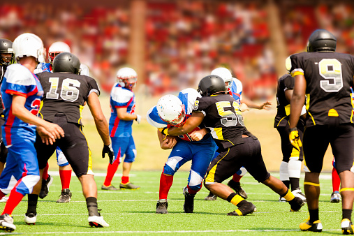 Semi-professional football team's running back carries the football to make a play. Defenders try to tackle him. Football field with a stadium full of unrecognizable fans in background.