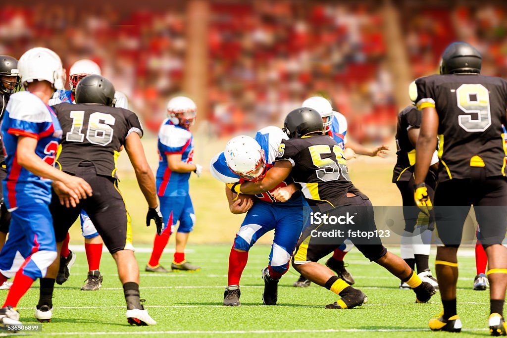 Football team's running back carries ball. Defenders. Stadium fans. Field. Semi-professional football team's running back carries the football to make a play. Defenders try to tackle him. Football field with a stadium full of unrecognizable fans in background. American Football - Sport Stock Photo