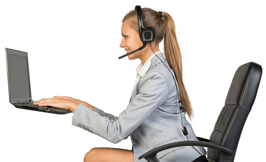 Businesswoman in headset sitting on office chair, typing on laptop with blank screen, smiling. Isolated over white background