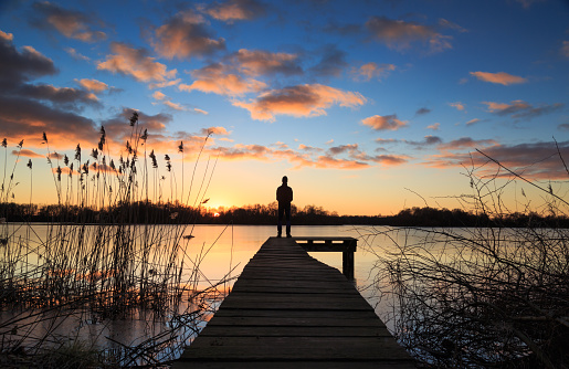 Man standing on a small jetty, enjoying the winter sunset over a lake.