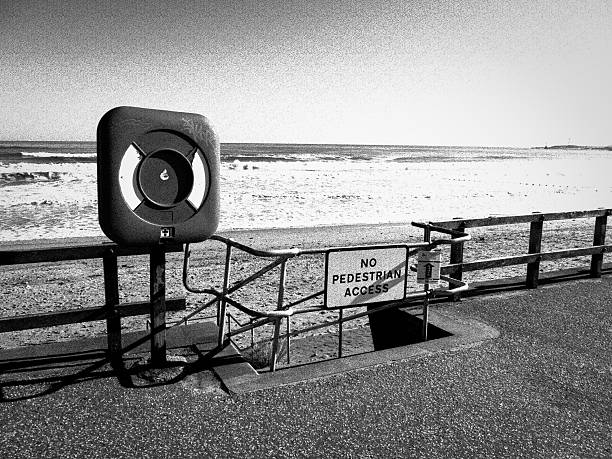 This grainy, Black and White Image show a Life Ring station on the pubic Beach Front at Aberdeen, UK.  On a stormy day, with the North Sea being churned up in the background, a 'No Pedestrian Access' sign, is placed in front of the steps leading to the beach, for safety reasons.
