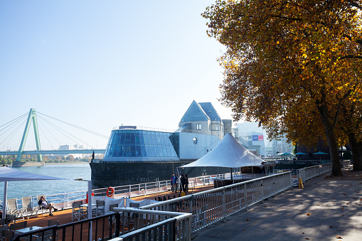 Cologne, Germany - October 27, 2015: Zollhafen with Hussel Confiserie and Imhoff Schokoladenmuseum at river Rhine seen from promenade Holzwerft in autumn. In foreground people are on deck of tourboat.