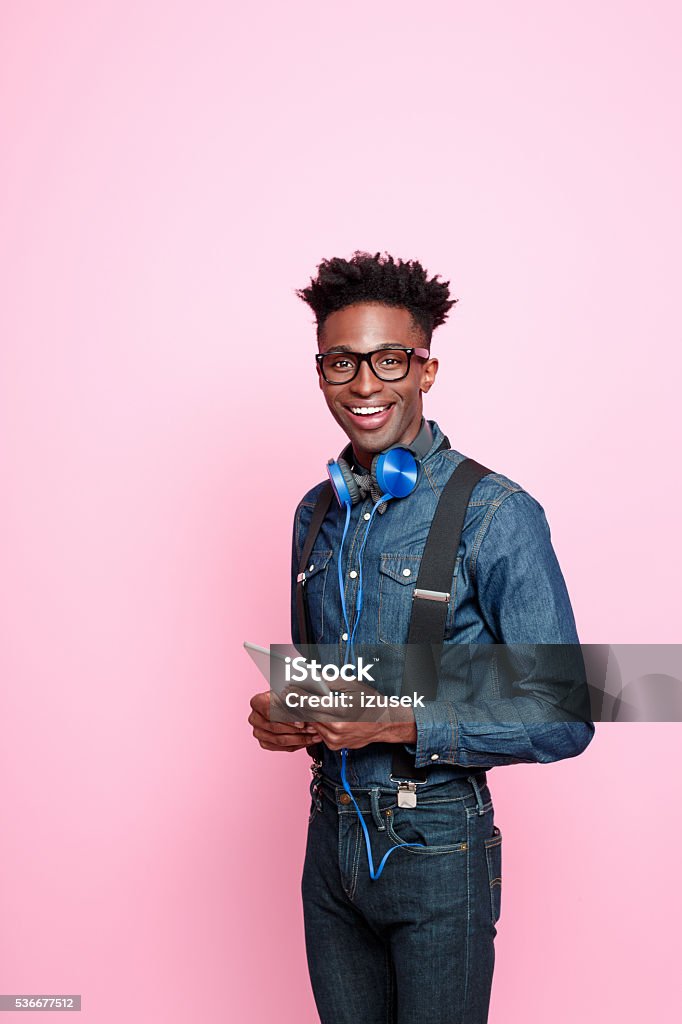 Friendly afro american guy in fashionable outfit holding digital tablet Studio portrait of friendly afro american young man wearing denim shirt and trausers and headphone, holding a digital tablet in hands and smiling at camera. Studio portrait, pink background. Men Stock Photo