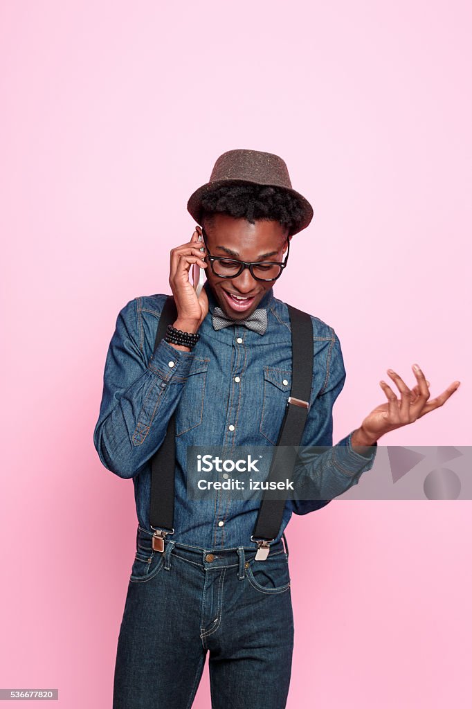 Funky afro american guy talking on smart phone Studio portrait of friendly afro american young man wearing denim shirt and trausers, hat, nerd glasses and headphone, talking on smart phone. Studio portrait, pink background. Discussion Stock Photo