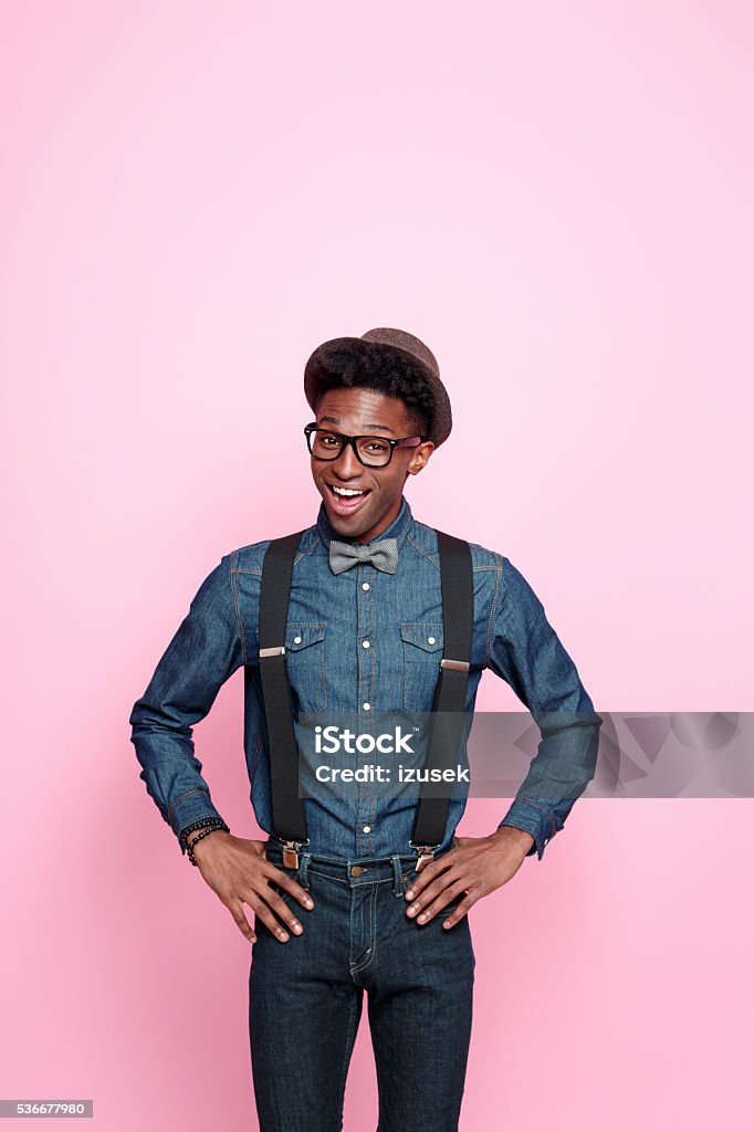 Portrait of fashionable afro american guy Studio portrait of friendly afro american young man wearing denim shirt and trausers, hat, nerd glasses and bowtie, smiling at camera. Studio portrait, pink background. Men Stock Photo
