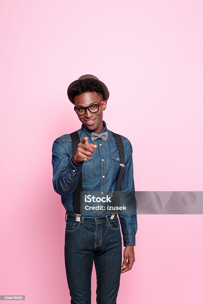 Fashionable afro american guy pointing with index finger Studio portrait of friendly afro american young man wearing denim shirt and trausers, hat, nerd glasses and bowtie, pointing with index finger at the camera. Studio portrait, pink background. Pointing Stock Photo