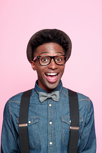 Studio portrait of excited afro american young man wearing denim shirt, hat, nerd glasses and bow tie. Studio portrait, pink background.