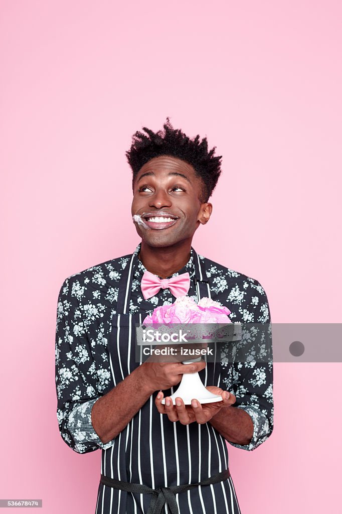 Cute afro american small business owner holding cookies Portrait of cute afro american young man wearing floral patern shirt, pink bow tie and apron, holding cookies. Studio shot, pink background. Baker - Occupation Stock Photo