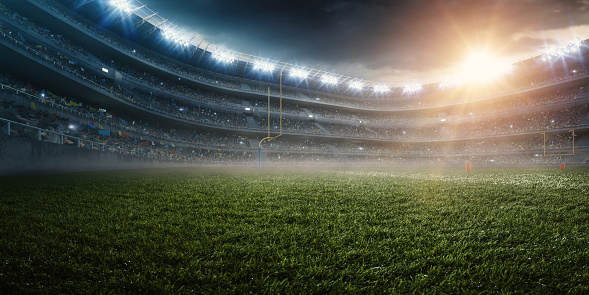 A wide angle panoramic image of a outdoor american football stadium full of spectators under evening sky. The image has depth of field with the focus on the foreground part of the pitch. The view from center of the field.