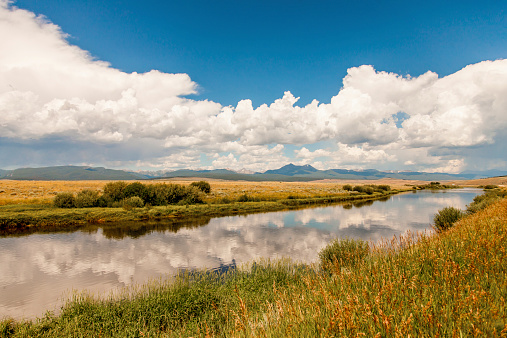 Montana landscape. Horizontal image shows a vast landscape of yellow and green foliage, alongside a reflective river. Blue sky and clouds above.