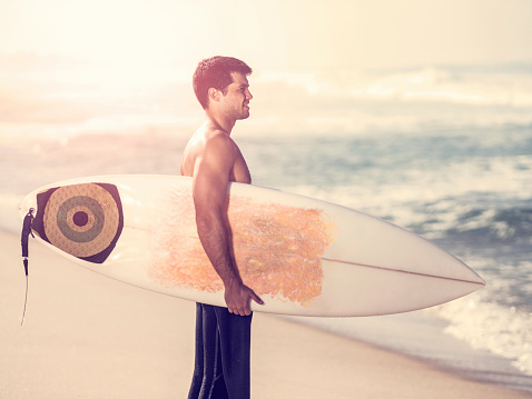 Side view of young Latin American surfer standing with surfboard at sunset.