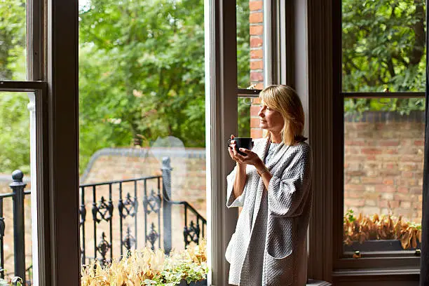 Shot of a mature woman drinking tea while looking out of her living room window