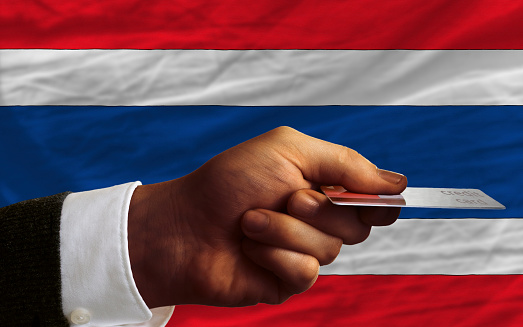 man stretching out credit card to buy goods in front of complete wavy national flag of thailand