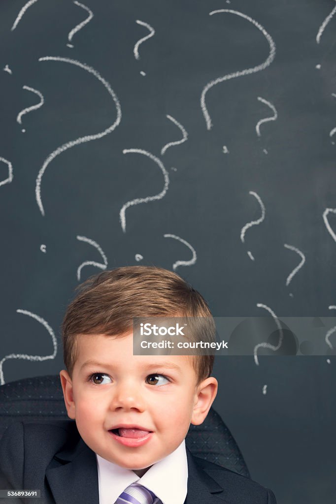 Humorous Decision making concept. Young child in a suit thinking Humorous Decision making concept. Young child in a suit thinking with question marks behind him on a black board. He is wearing a suit and looking like he has found a solution. 2-3 Years Stock Photo