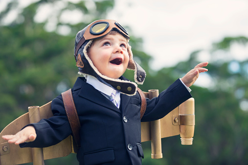 Aspiration, innovation business concept. A young smiling child wearing home made costume of a traditional pilots outfit complete with eye goggles and cap with ear flaps. A blurry background of natural greenery places the child in sharp focus, allowing for focus on the cardboard plane strapped into his back.