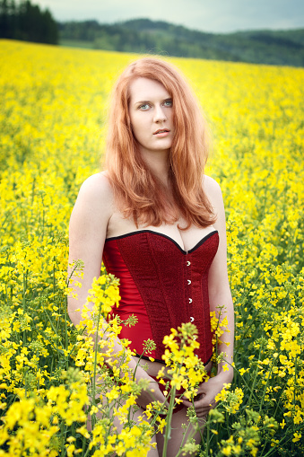 Beautiful redhead in a red corset posing in the oilseed Rape, in a nature. Glamour and nude photography.