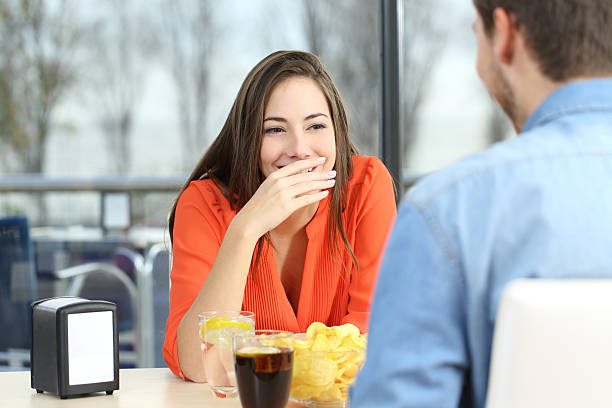 Woman covering her mouth to hide smile or breath Woman covering her mouth to hide smile or bad breath during a date in a coffee shop with a window in the background couple on bad date stock pictures, royalty-free photos & images