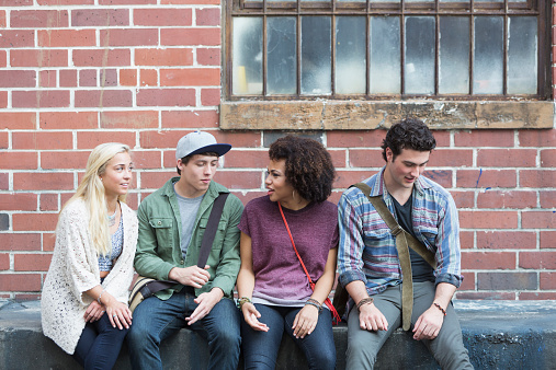 A group of four multiracial young adults hanging out together.  They are sitting side by side outside an old brick building with security bars on the window.  They are casually dressed in jeans.