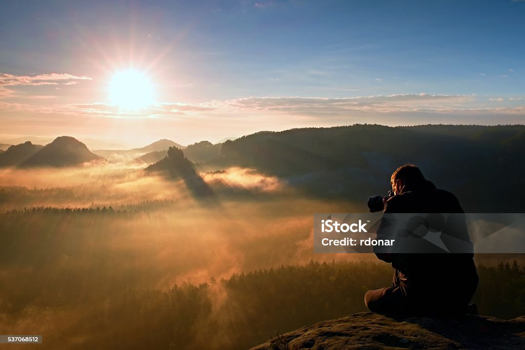 Happy tourist with camera watching into colorful misty  morning Tourist sit on the peak of sandstone rock and watching into colorful mist and fog in morning valley. Adult Stock Photo