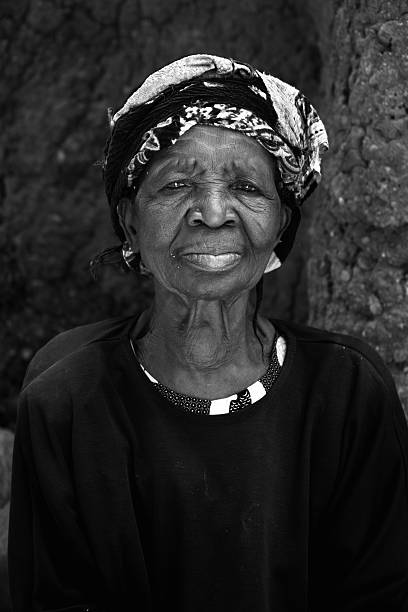 An elderly woman from a village in northern Togo, West Africa wearing a head scarf and with a traditional cob wall behind her.