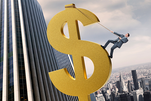 A businessman using a rope to climb to the top of a large dollar sign surrounded by skyscrapers and the New York City skyline in the distance. Standing on a giant gold dollar sign, the business man has lassoed capital, using money to make money. Money is important to him and he scaling the income ladder. 