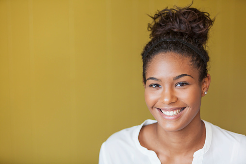 Portrait of beautiful African American teenage girl.  She is smiling at the camera.  Her hair is pulled back into a bun.  The background is a dull yellow wall.