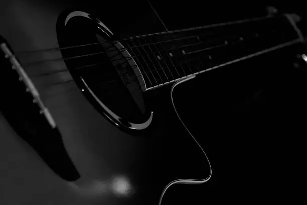 A close up black and white DSLR photo of an Yamaha APX electro acoustic guitar in dark and blur background. The guitar is standing free on a table in a dark room at night, artificial flash light with snoot is coming from front end and spread accross the guitar neck. Background is all black.