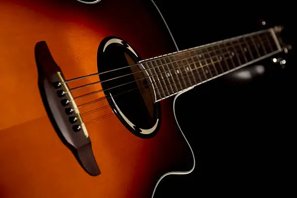 A close up DSLR photo of an rosewood, red electro acoustic guitar in dark and blur background. The guitar is standing free on a table in a dark room at night, artificial flash light with snoot is coming from front end and spread accross the guitar neck.