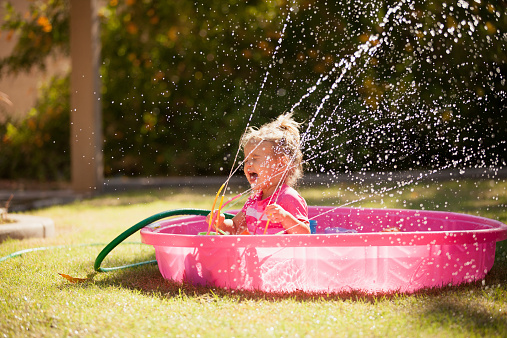 Little girl splashing in a pink kiddie pool with the hose and sprinkler spraying in her face.