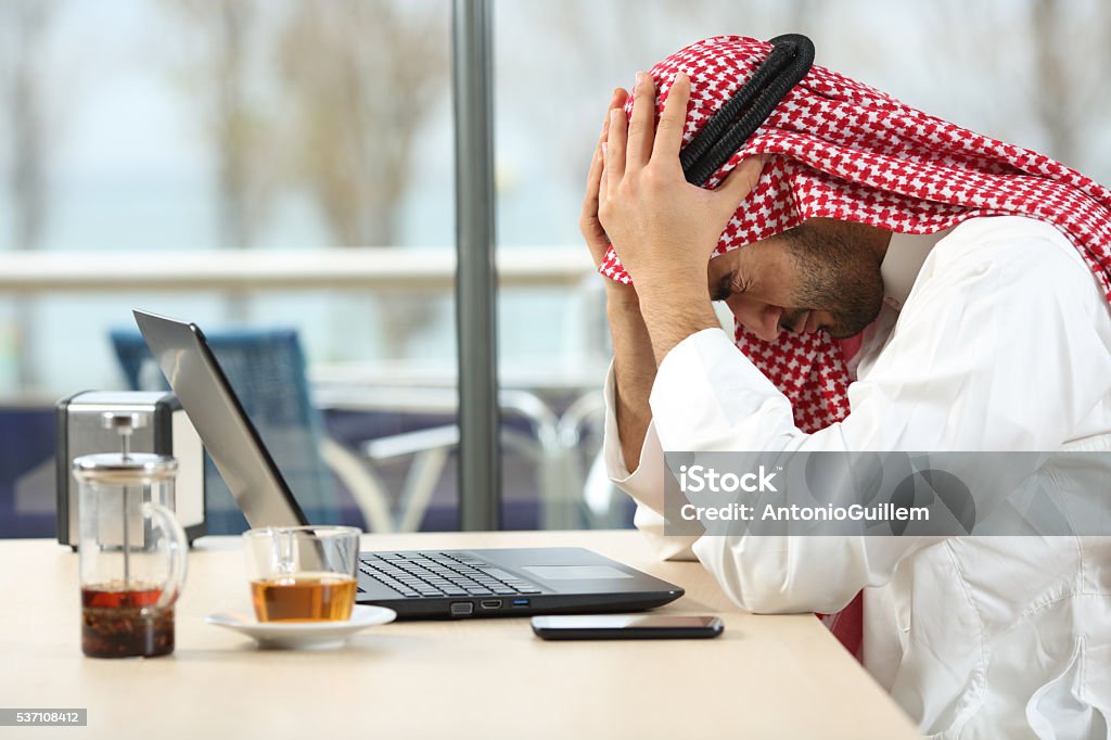 Desperate arab saudi man online bankruptcy Profile of a desperate and alone arab saudi man with a laptop online in a coffee shop with a window in the background. Bankruptcy concept Men Stock Photo