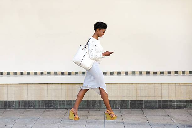молодая женщина, ходьба и отправив текстовое сообщение на мобильный телефон - smart casual women full length casual стоковые фото и изображения