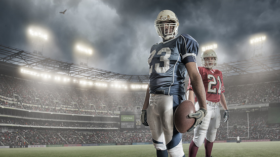 Portrait of two American football players from rival teams, standing and holding ball before a game . Set in a generic outdoor floodlit stadium full of spectators under dramatic stormy sky. Both players are wearing unbranded generic kit. This is a composite image created in Photoshop with fake stadium advertising. 