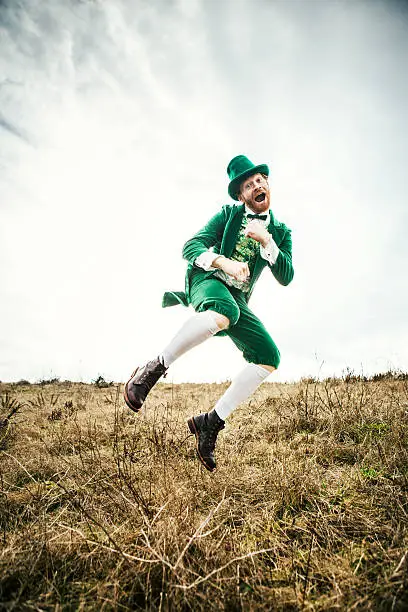 A stereotypical Irish character all ready for Saint Patricks day jumps and dances in an open field of Irish country side.  Copy space in the sky and grass.