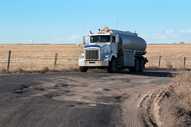 A flammable liquids semi truck drives over potholes and washboard which lines county road 127 next to the Pawnee National Grasslands and runs through the Niobrara oil fields. A DOT flammable liquids sign number 3 -1203 for gasoline sits on top and sides of the truck. Roads are damaged by the increased heavy petroleum industry traffic in the area which runs day and night. Oil storage tanks stand on the horizon in the background.