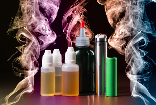 Bottles of E-juice next to a Rebuildable dripping atomizer surrounded by colored smoke 