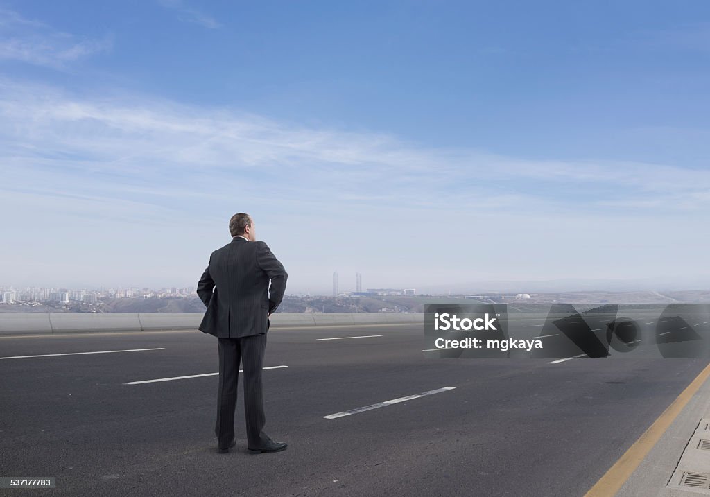 Businessman on the Highway Rear view of businessman standing and looking on the highway with city and mountainous landscape view background. He is looking further, thinking and searching new ideas. Highway Stock Photo