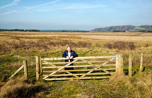 Middle age man standing behind  a large wooden gate to an open marsh like field